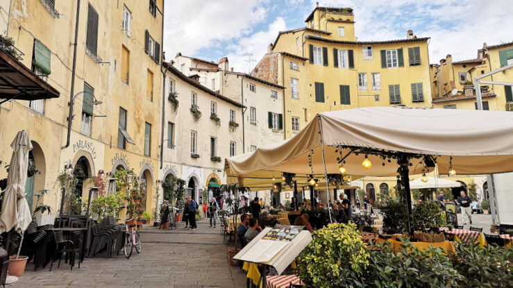 Outdoor cafés beneath large umbrellas in Piazza dell'Anfiteatro, surrounded by colourful historic buildings in Lucca's medieval centre, Tuscany, Italy